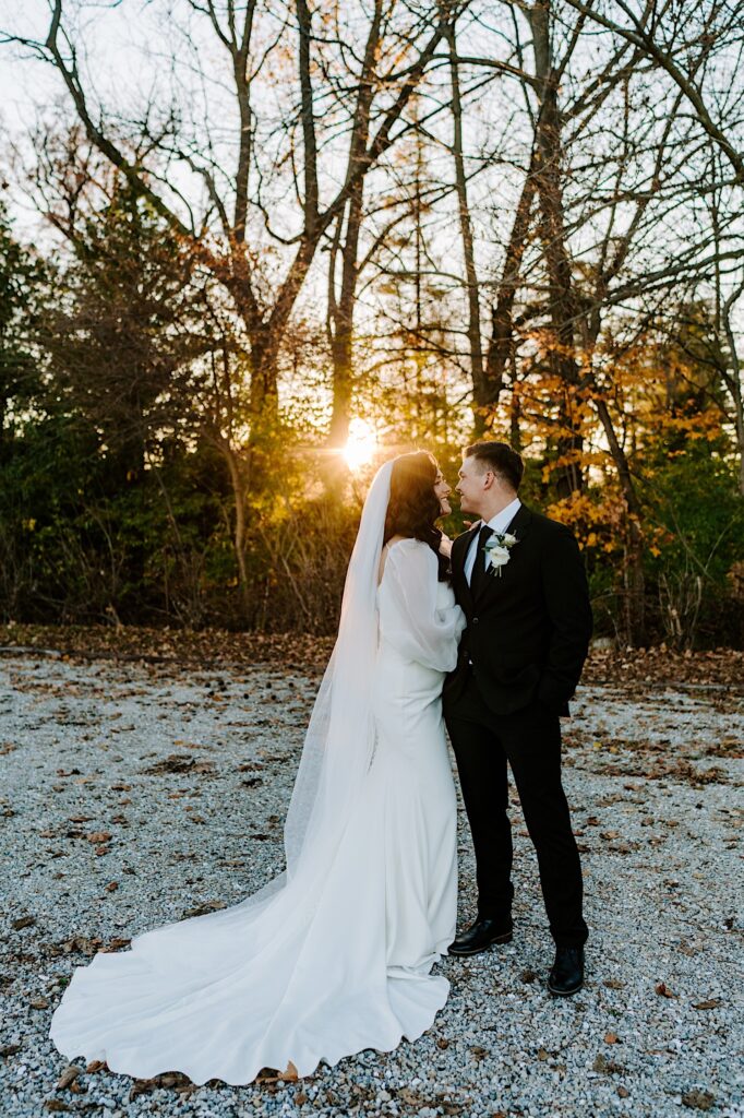 A bride and groom smile at one another while the sun sets behind them on the forest