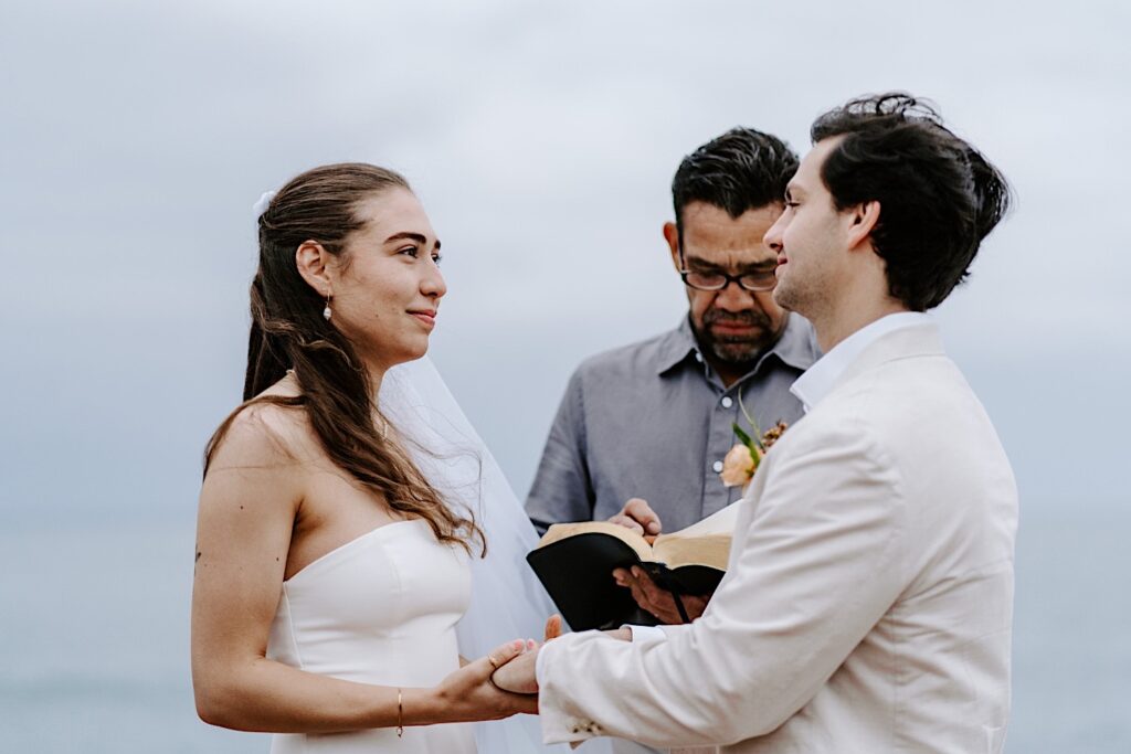 A bride and groom hold hands and smile at one another while their officiant reads during their destination elopement ceremony in San Diego