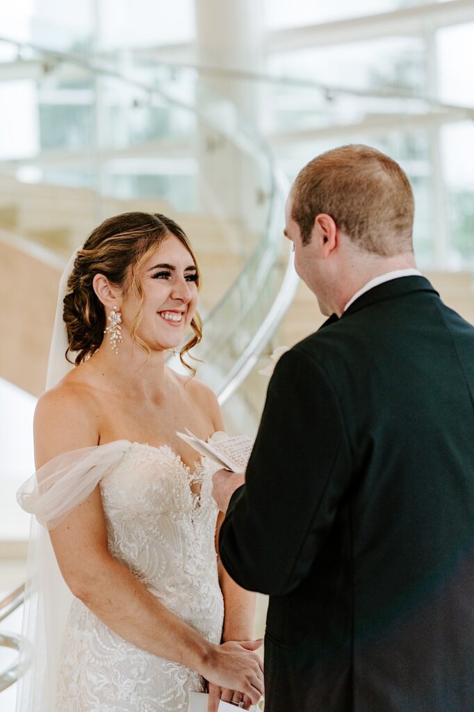 A bride smiles while the groom reads his private vows to her while they stand together in the lobby of their hotel