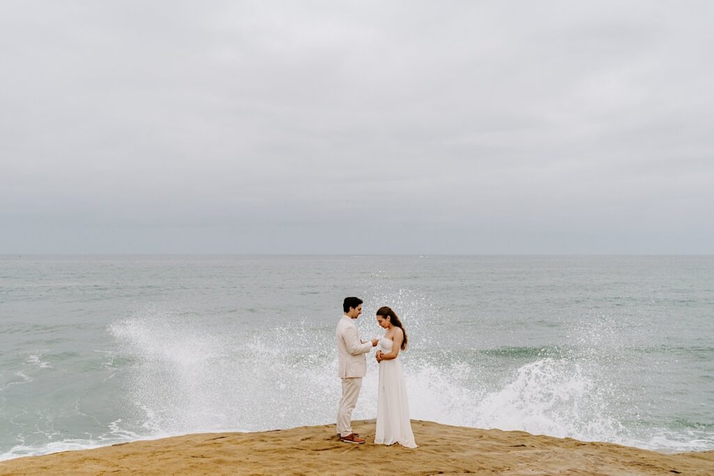 A bride and groom stand facing one another on a cliff as waves of the ocean in the background crash near them, photo taken by a destination wedding photographer