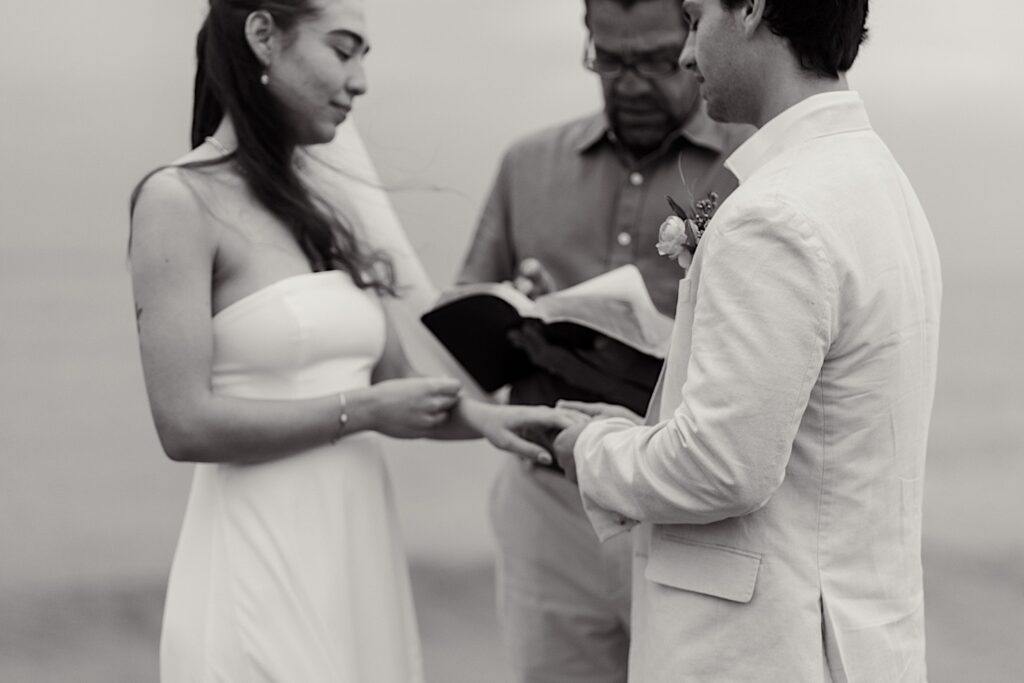 Black and white photo of a groom putting the wedding ring on the bride's hand during the ceremony of their destination elopement in San Diego