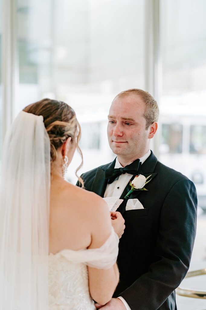 A groom tears up as the bride stands in front of him and reads her vows while they stand in the lobby of their hotel