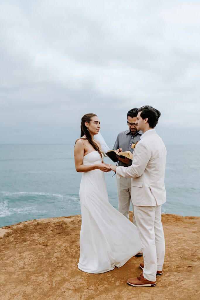 A bride puts the wedding ring on the hand of the groom during their elopement ceremony on the cliffs in San Diego
