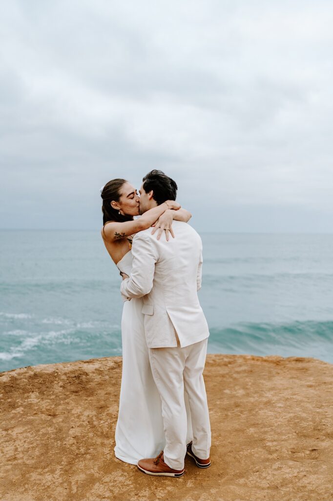 A bride and groom kiss one another while standing on the cliffs in San Diego on a cloudy day