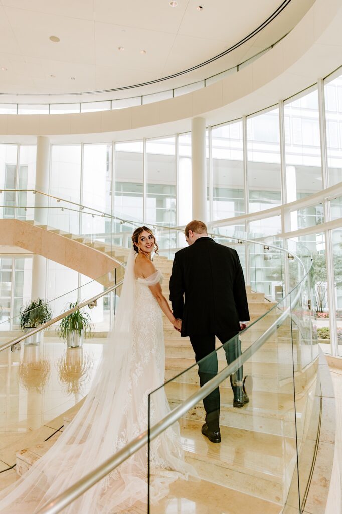 A bride looks over her shoulder as her and the groom walk up a set of stairs in the lobby of their hotel