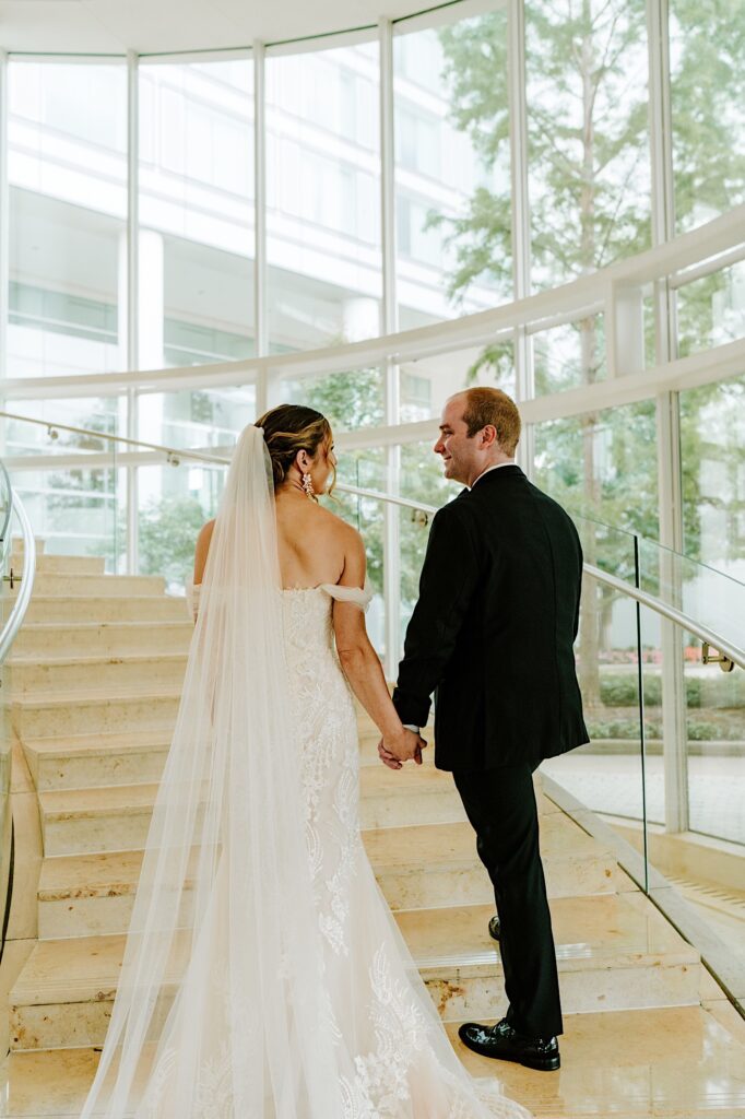 A bride and groom look at one another while holding hands and walking up a set of stairs in the lobby of their hotel
