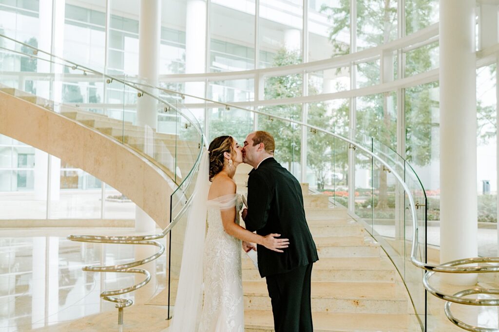 A bride and groom kiss one another while standing in front of a set of stairs in the lobby of their hotel
