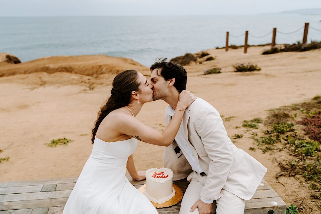 A bride and groom kiss one another with their wedding cake in between them while on a bench at the cliffs in San Diego for their destination elopement