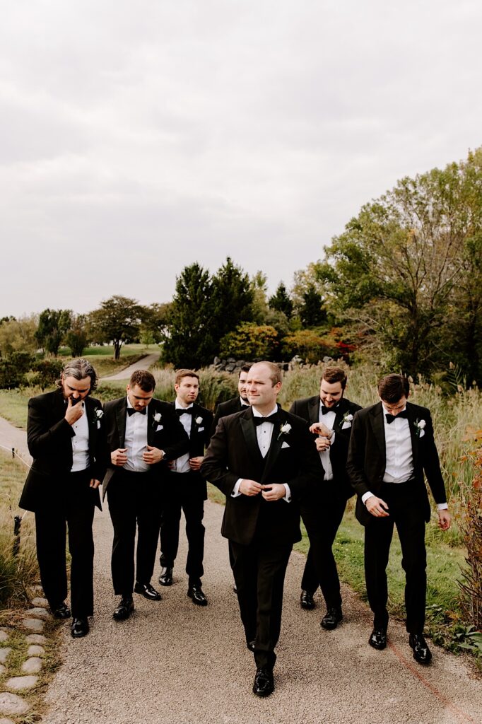 A groom walks and adjusts his suit coat while his 6 groomsmen walk behind him at the Makray Golf Club