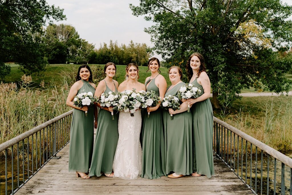 A bride and her 5 bridesmaids stand on a wooden bridge at the Makray Golf Club and smile at the camera before the bride's Chicagoland wedding