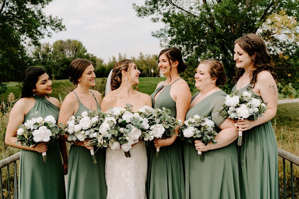 A bride smiles at her 5 bridesmaids around her who all smile back on the grounds of the Makray Golf Club before her wedding in the Chicagoland area