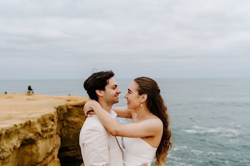 A bride and groom smile at one another as they embrace on the cliffs of San Diego after the ceremony of their destination elopement