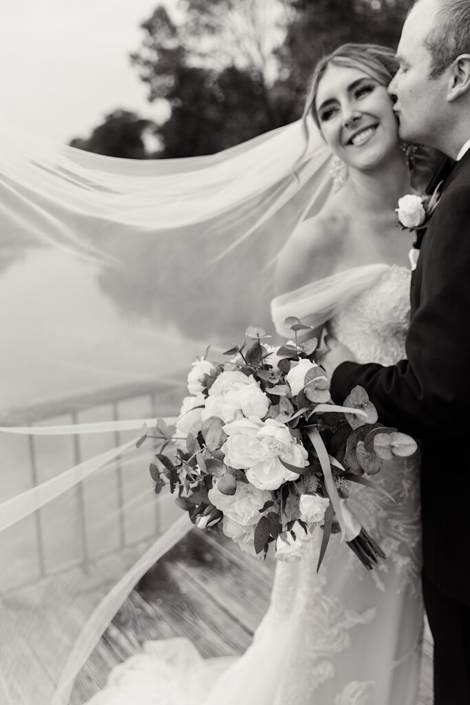 Black and white photo of a bride smiling as the groom kisses her on the cheek before their wedding at the Makray Golf Club