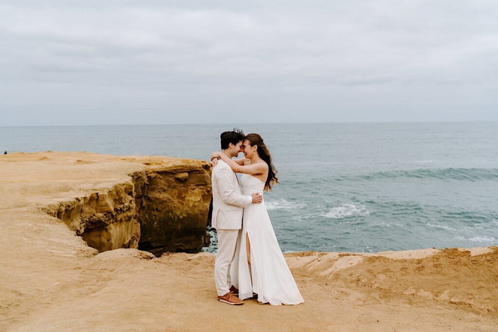 A bride and groom smile at one another and embrace while standing on the cliffs in San Diego after the ceremony of their destination elopement