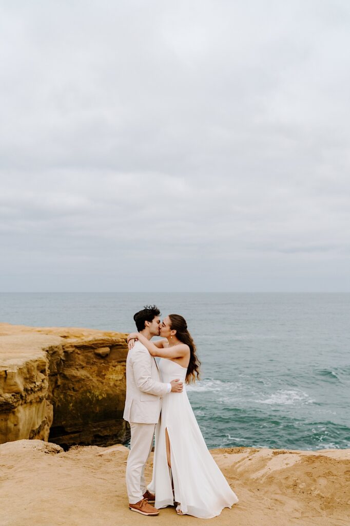 A bride and groom kiss one another while standing on the cliffs of San Diego on a cloudy day