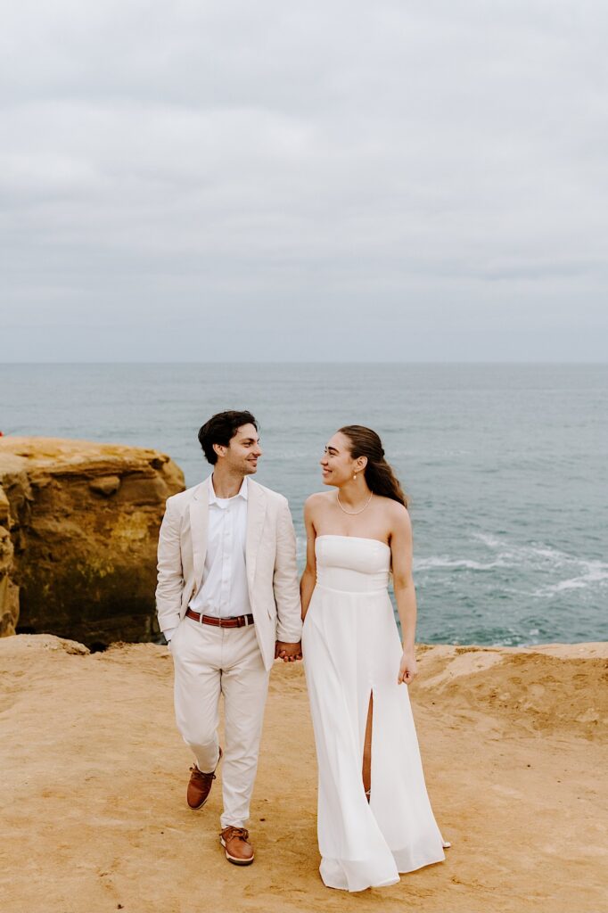 A bride and groom hold hands and smile at one another while walking towards the camera at the cliffs in San Diego