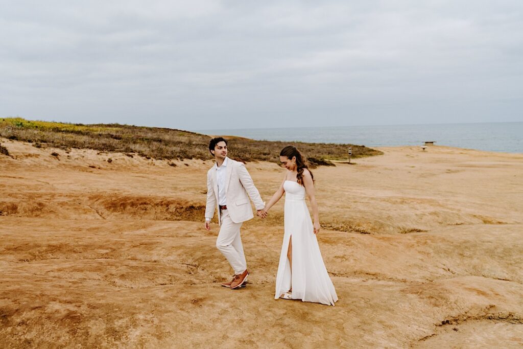 A bride and groom hold hands while walking along the cliffs in San Diego after the ceremony of their destination elopement