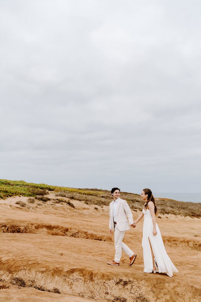 A bride and groom hold hands while walking along a beach in San Diego on a cloudy day