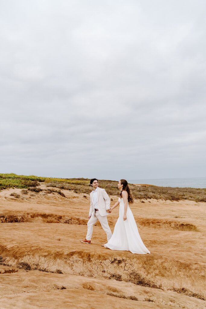 A bride and groom hold hands while walking along a beach in San Diego on a cloudy day