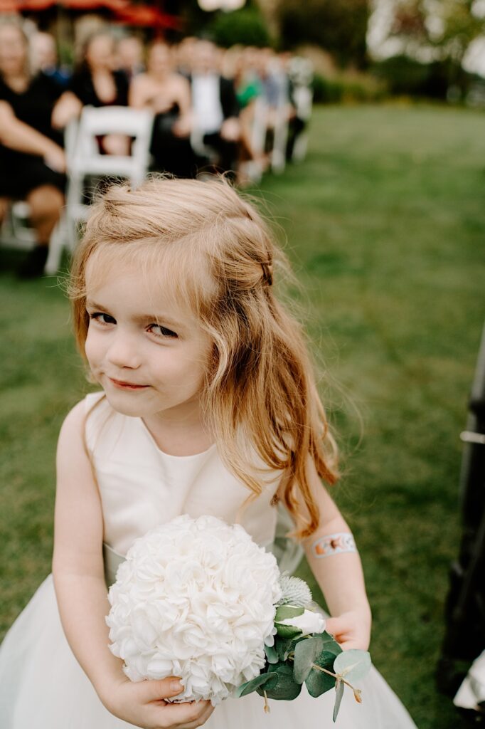 A flower girl gives a smirk to the camera while walking down the aisle of an outdoor wedding ceremony at the Makray Golf Club