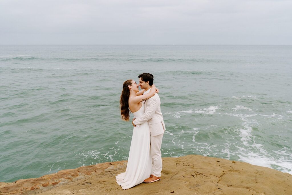 A bride and groom embrace and smile as they are about to kiss while standing on a cliff in San Diego after the ceremony of their destination elopement