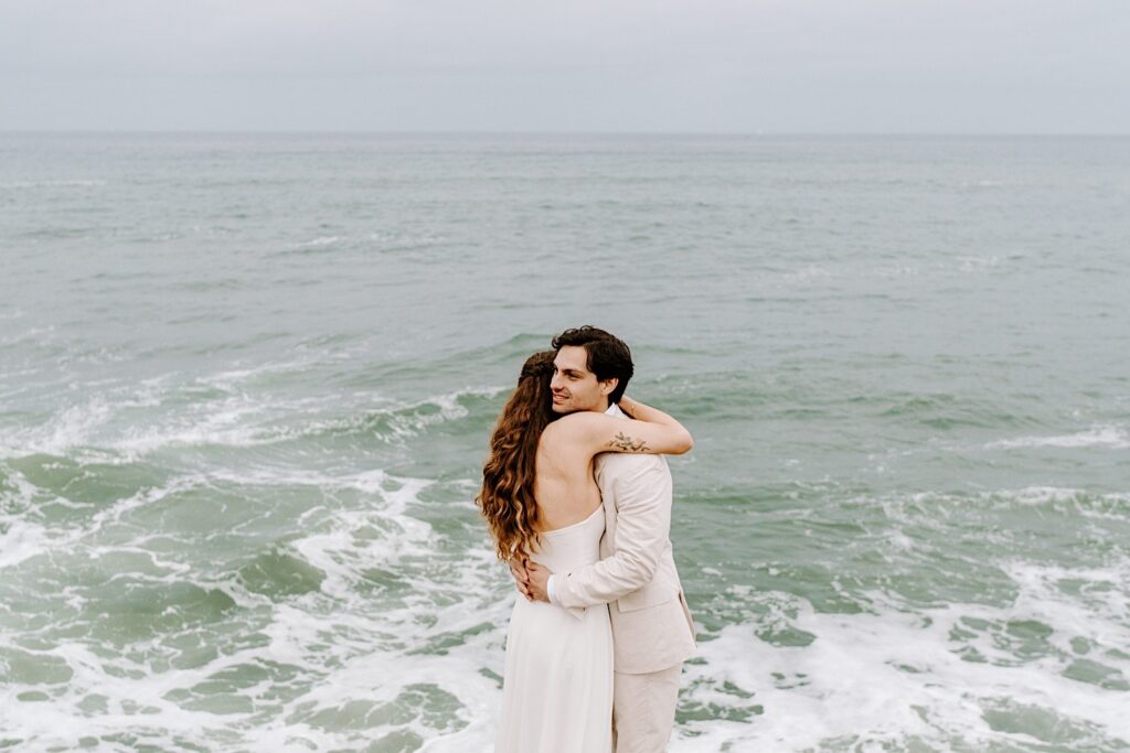 A groom smiles while hugging the bride next to the ocean as the two celebrate their destination elopement in San Diego