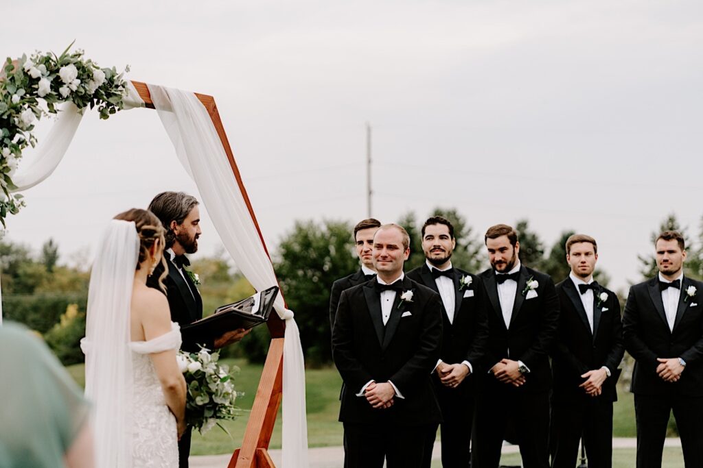 A groom stands next to his groomsmen while looking at the bride during their outdoor wedding ceremony at the Makray Golf Club in the Chicagoland area