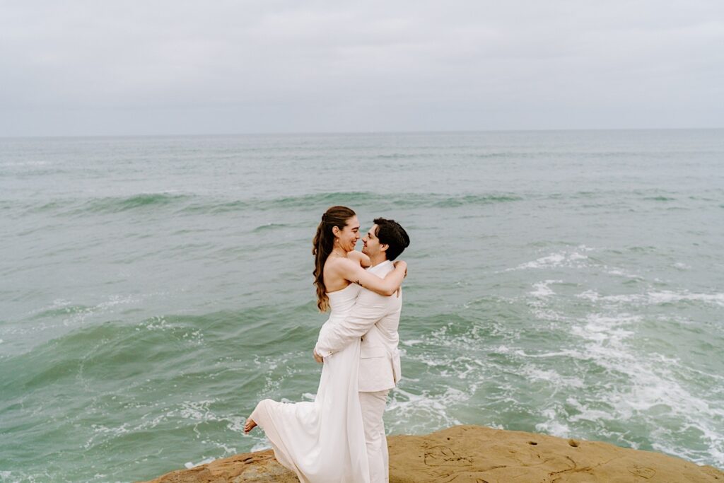 A bride smiles as the groom lifts her in the air while the two stand on a cliff in San Diego after the ceremony of their destination elopement