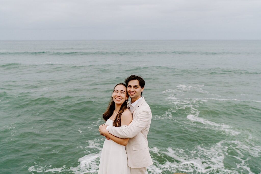 A bride and groom smile at the camera as the groom hugs the bride from behind while they stand in front of the ocean after their destination elopement in San Diego