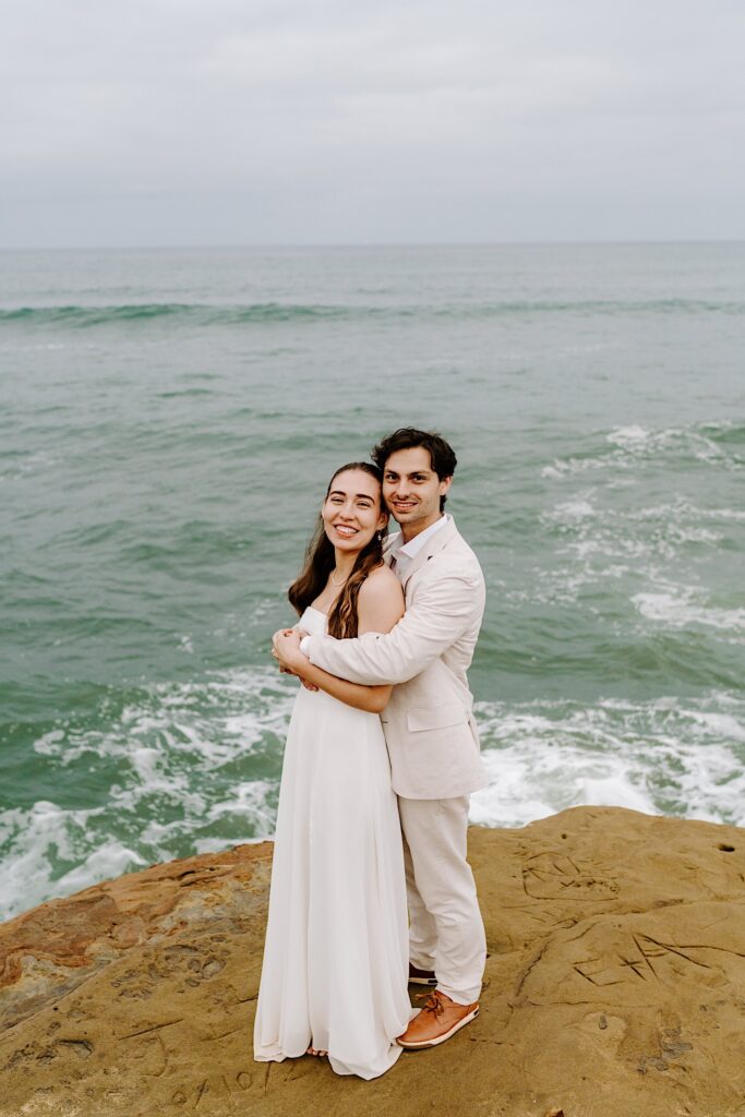 A bride and groom smile at the camera while standing on a cliff in San Diego with the ocean behind them