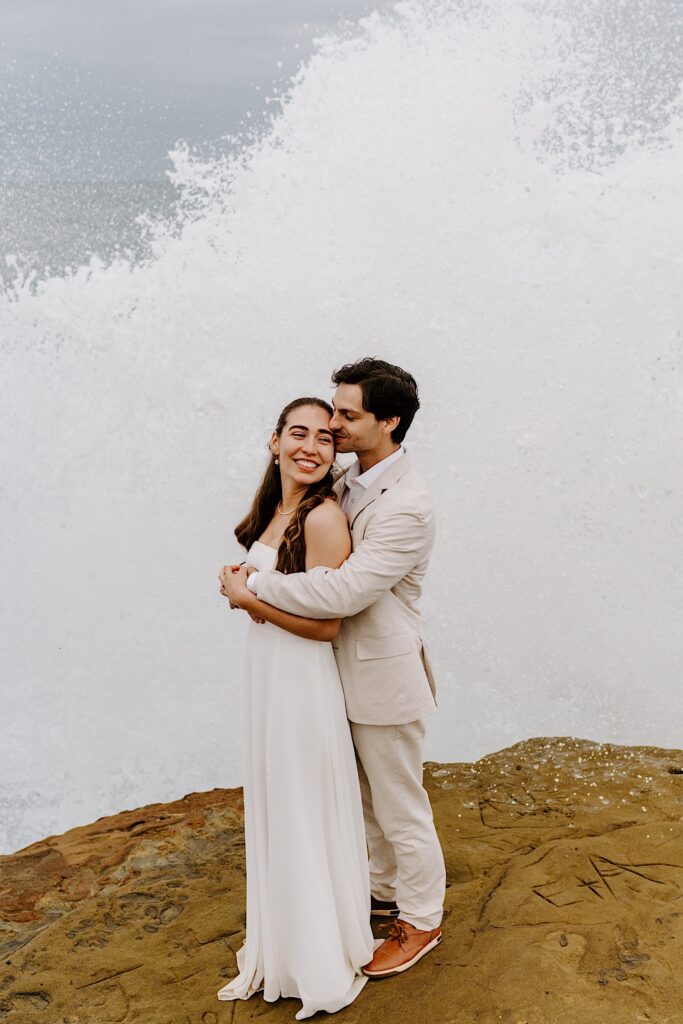 A bride smiles while the groom hugs her from behind and kisses her cheek on the cliffs in San Diego as the waves crash behind them