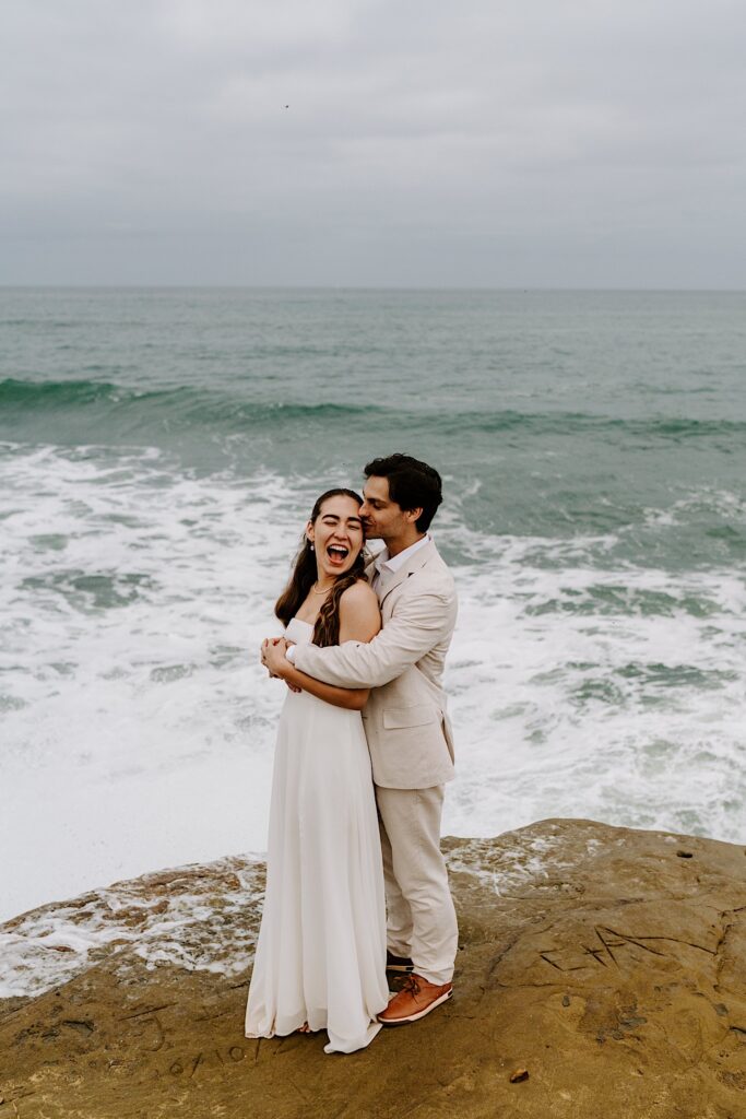 A bride exclaims after getting wet from a wave on the cliffs in Sand Diego as the groom hugs her from behind