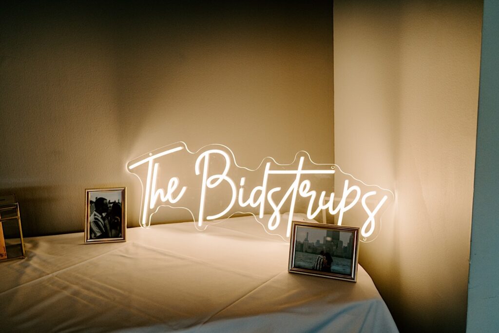 A neon sign of a bride and groom's last name is lit up on a table next to photos of them for the reception of their wedding