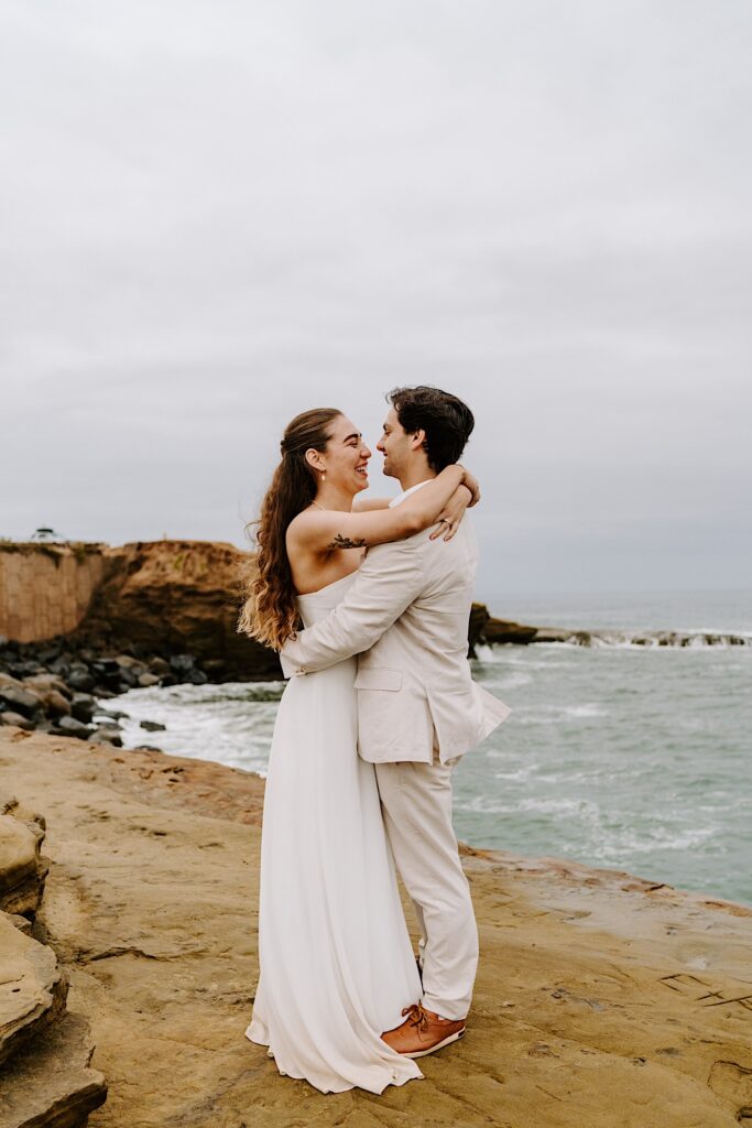 A bride and groom smile at one another while hugging on a beach in San Diego on a cloudy day