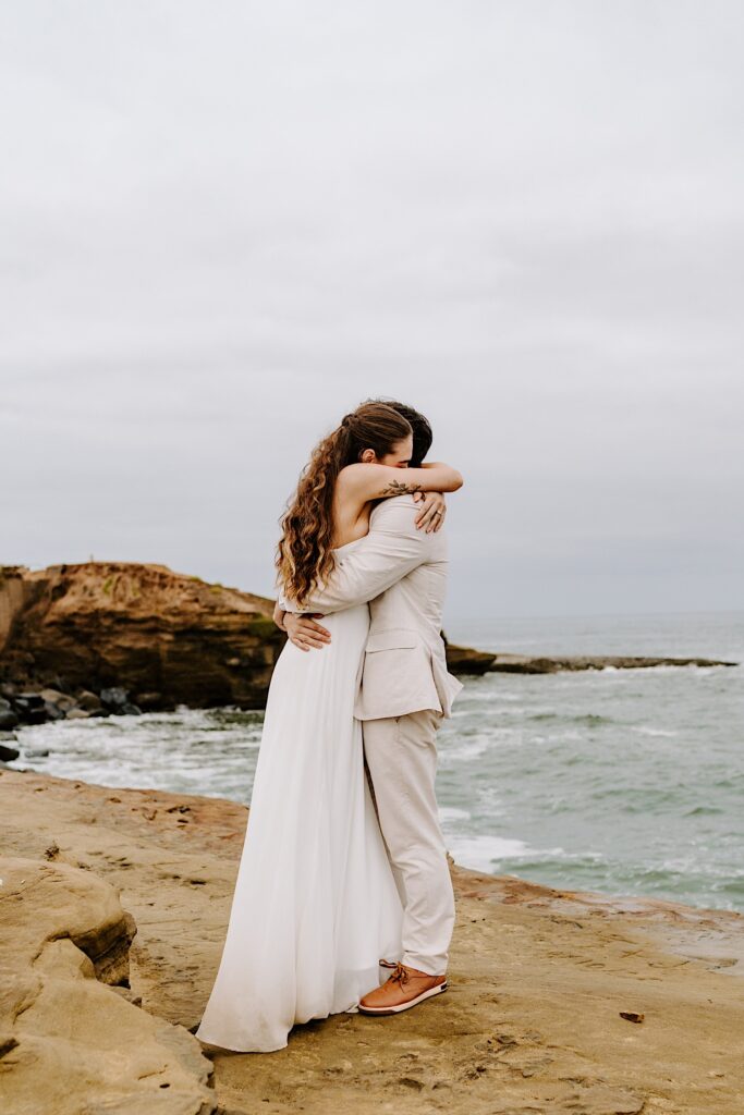 A bride and groom hug one another while on a beach in San Diego on a cloudy day
