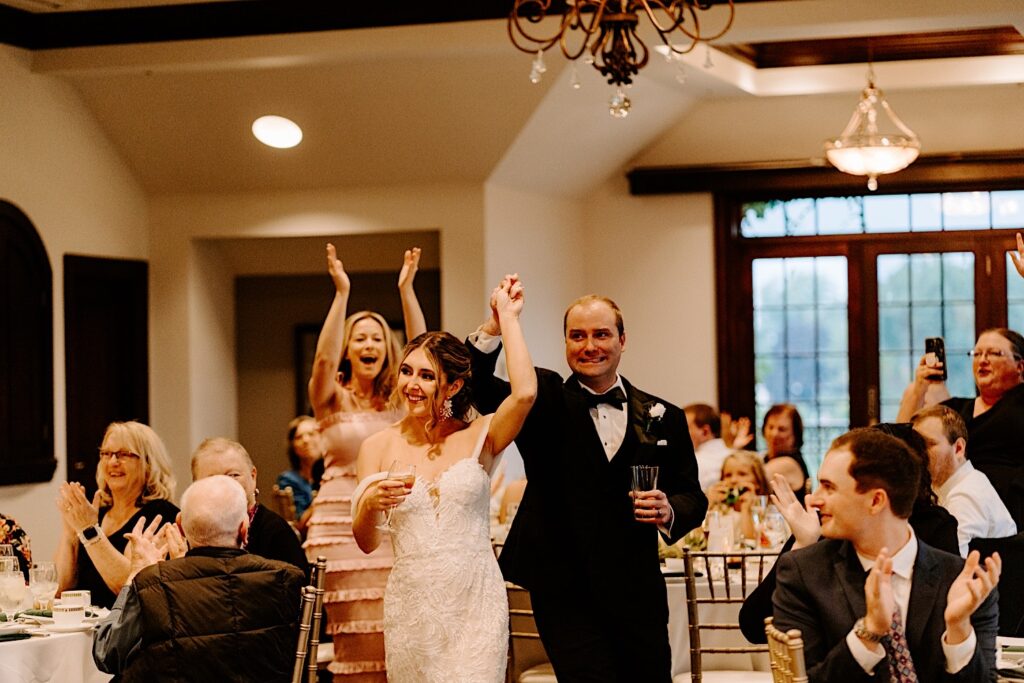 A bride and groom smile and raise their arms as they walk into their indoor wedding reception at the Makray Golf Club in the Chicagoland area, guests around them clap and cheer