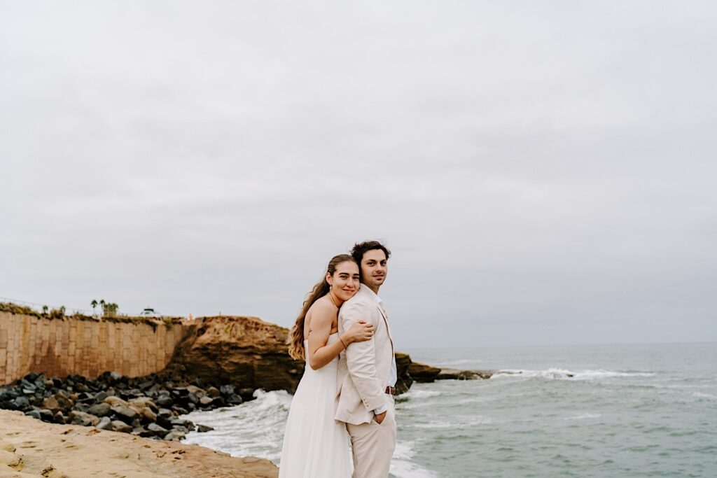 A bride hugs the groom from behind while the two smile at the camera on a beach in San Diego as they celebrate their destination elopement
