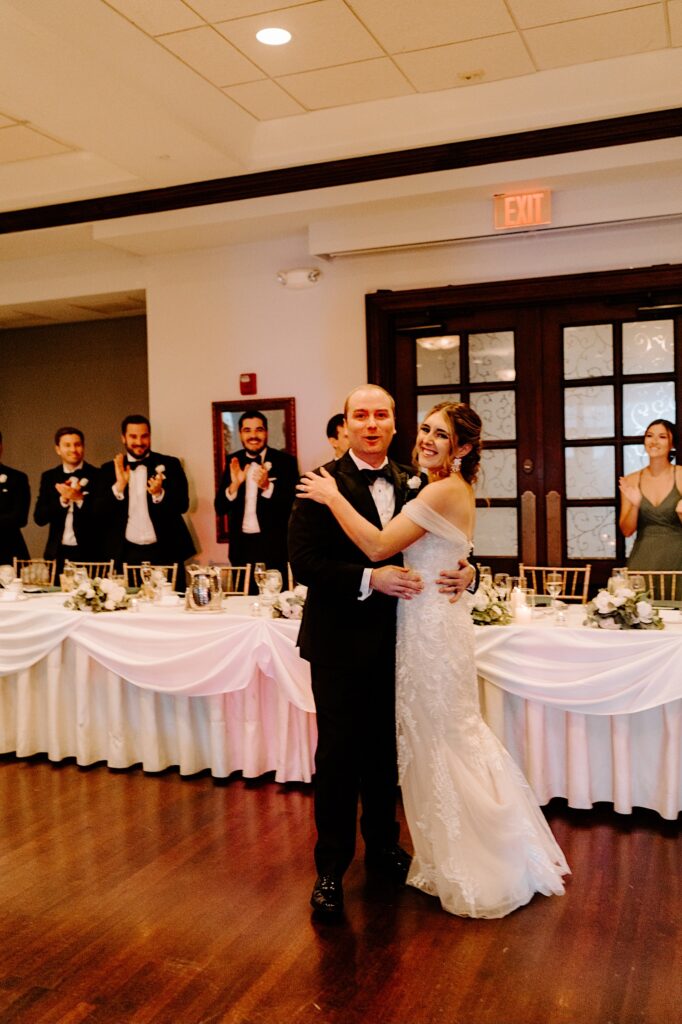 A bride and groom stand in front of their table during their indoor wedding reception and embrace one another