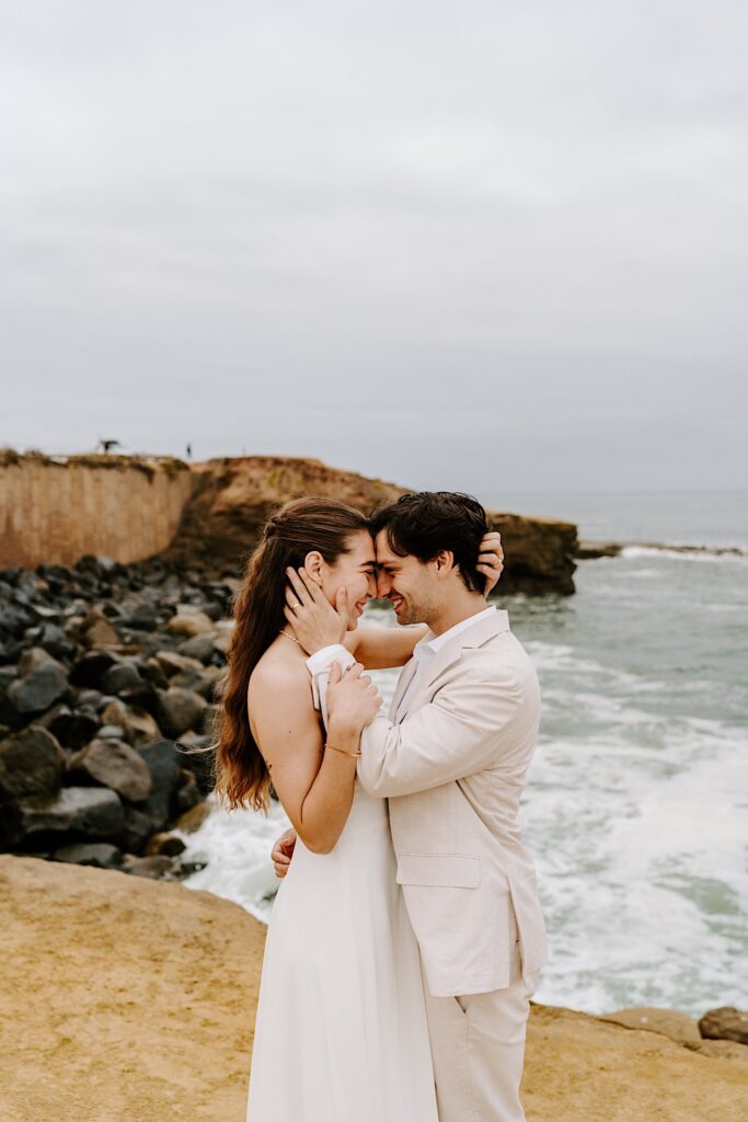 a bride and groom embrace and smile at one another while on a beach in San Diego on a cloudy day