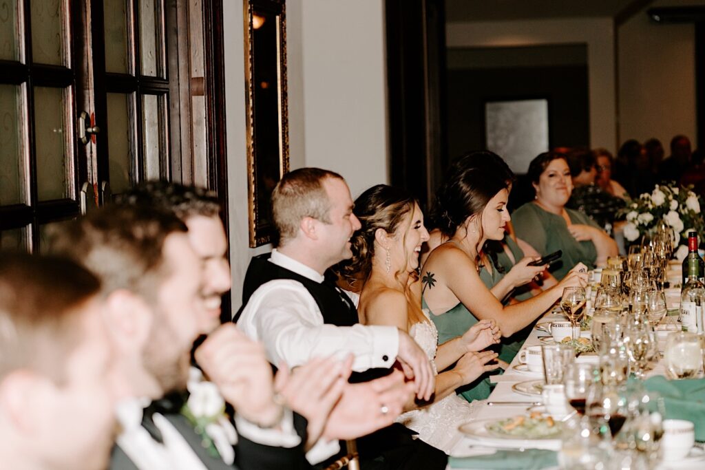 A bride and groom laugh while sitting next tot their wedding party members during their indoor reception at the Makray Golf Club in the Chicagoland area