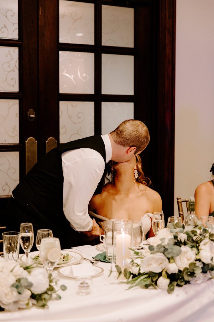 a groom stands and kisses the bride while she sits at their table during their indoor reception at the Makray Golf Club