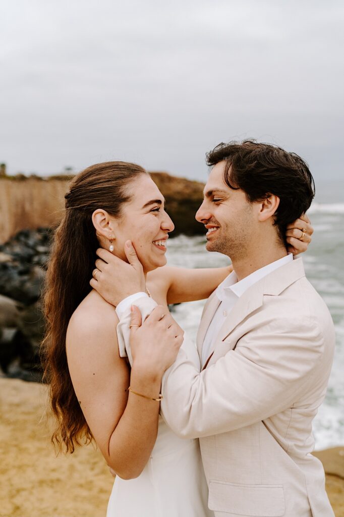 A bride and groom smile at one another as they embrace while  on a beach in San Diego on a cloudy day