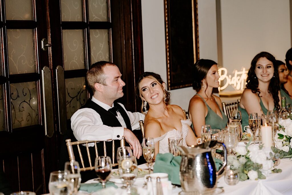 A bride smiles at the groom while sitting next to him at their table during their indoor wedding reception at the Makray Golf Club in the Chicagoland area