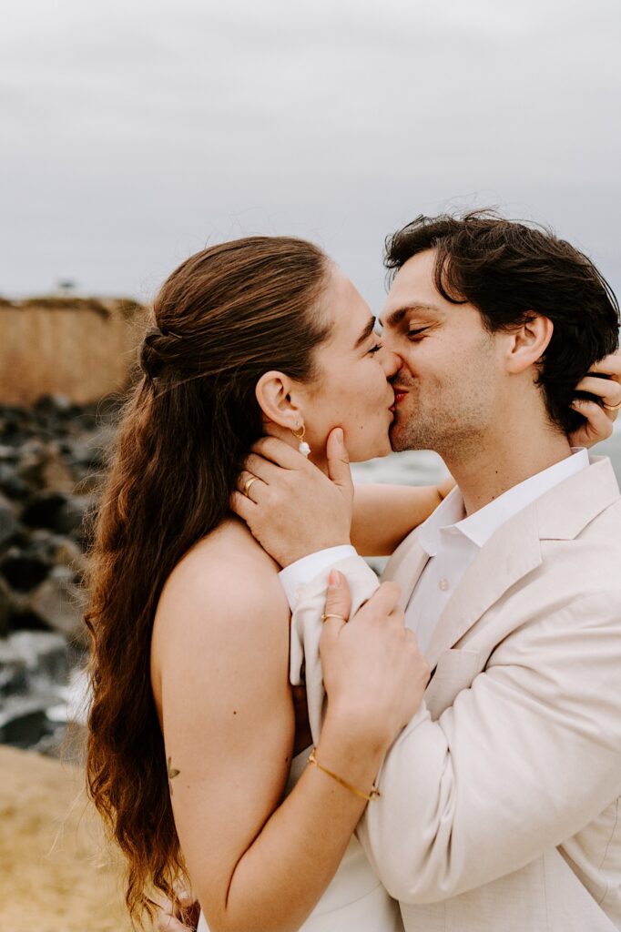 A bride and groom smile and kiss one another while on a beach in San Diego on a cloudy day