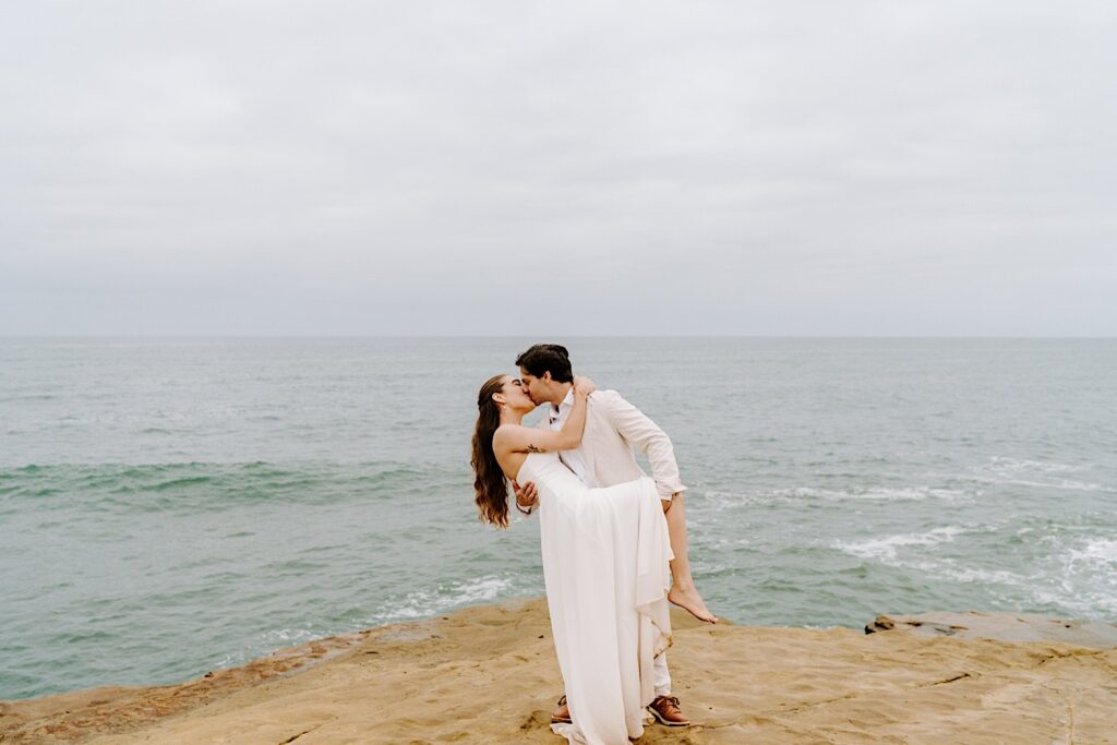 A bride and groom kiss while standing on the cliffs in San Diego as they celebrate their destination elopement