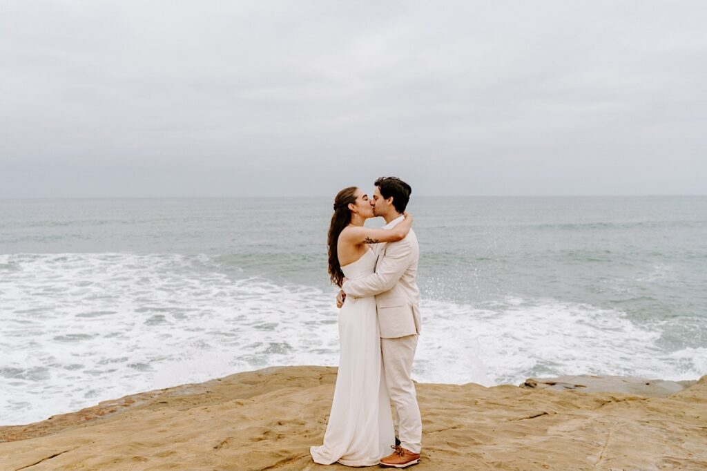 A bride and groom kiss one another as they embrace on the cliffs in San Diego during their destination elopement