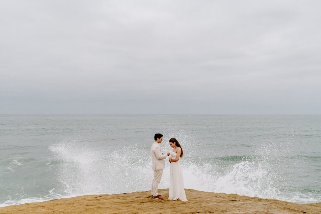 A bride holds her hands while looking down as the groom reads his private vows to her while on the cliffs in San Diego for their destination elopement