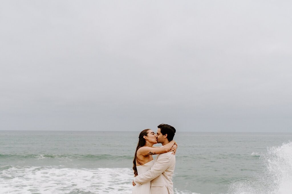 A bride and groom kiss one another while hugging in front of the ocean on the cliffs in San Diego during their destination elopement