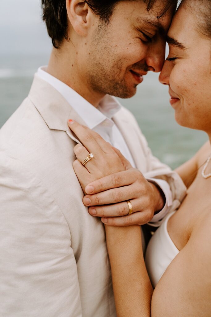 A bride and groom embrace and show off their wedding rings on the groom's chest while closing their eyes