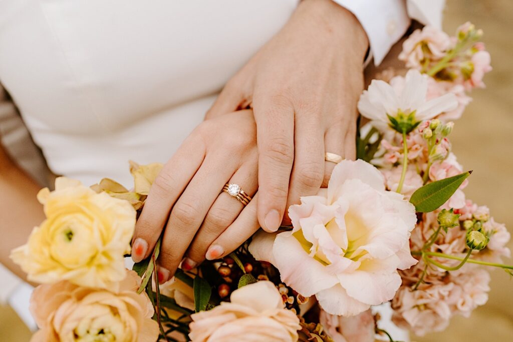 Close up photo of a bride and groom's hands with their wedding rings on them resting atop one another and holding a flower bouquet